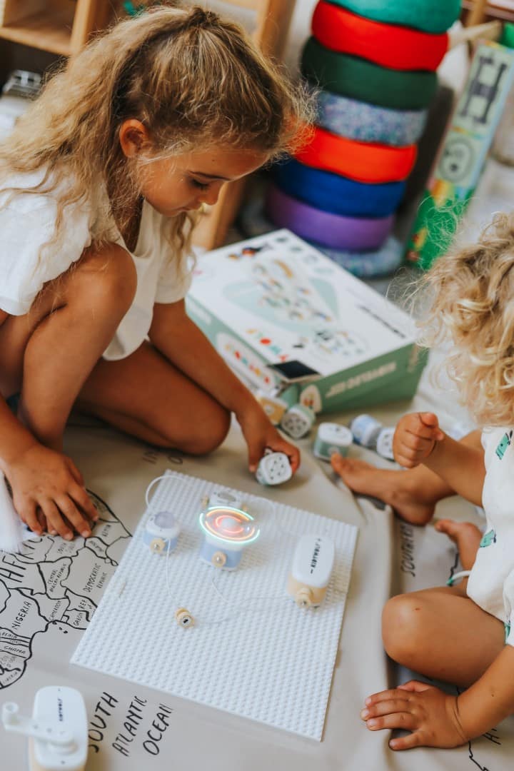 Criança de cabelo comprido a interagir com os componentes do kit KIDYELEC sobre um tapete de mapa, enquanto outra criança observa.