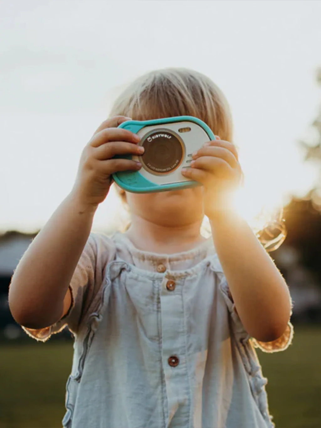 Criança loira a usar a máquina fotográfica digital Kidywolf à prova de água, tirando uma foto ao ar livre ao pôr do sol.