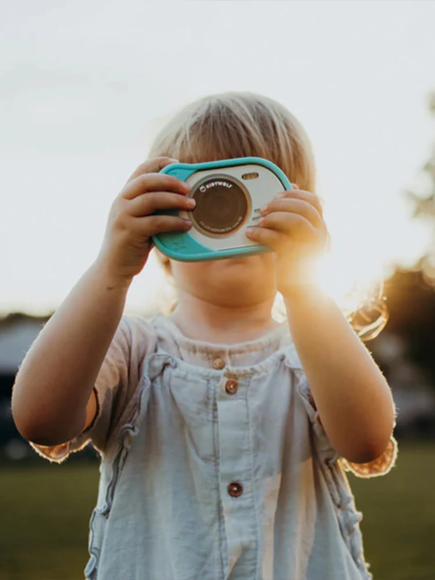 Criança loira a usar a máquina fotográfica digital Kidywolf à prova de água, tirando uma foto ao ar livre ao pôr do sol.