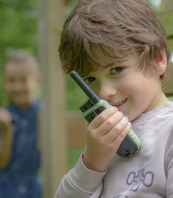 Criança sorridente a usar um walkie talkie KidyWolf verde e preto ao ar livre, em brincadeira com outra criança ao fundo.