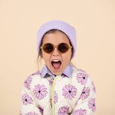 Criança com gorro lilás e casaco estampado, a usar os óculos de sol Kietla castanhos com expressão divertida.