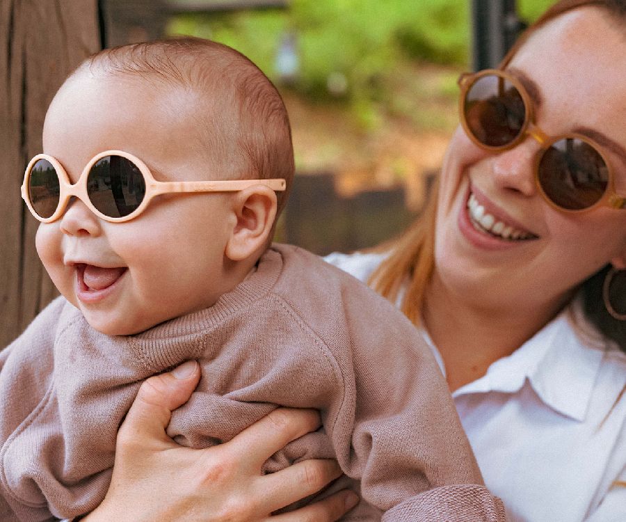 Bebé sorridente de 0-1 ano a usar óculos de sol Kietla Diabola cor pêssego, enquanto é segurado por uma mulher sorridente.