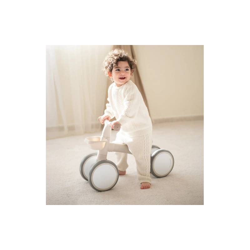 Criança sorridente de 1 a 3 anos a usar a bicicleta de equilíbrio Kikkaboo ZIPI de 4 rodas num ambiente interior claro.