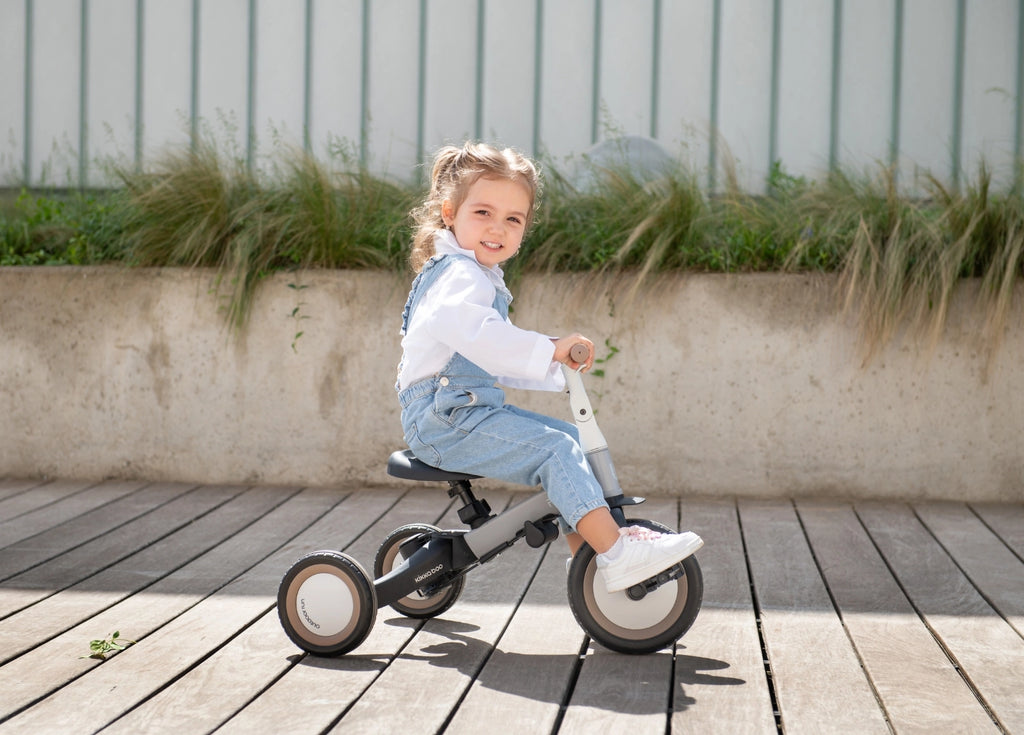 Criança sorridente de 3-4 anos a andar no triciclo evolutivo Kikkaboo FLIP bege e preto num deck de madeira exterior.