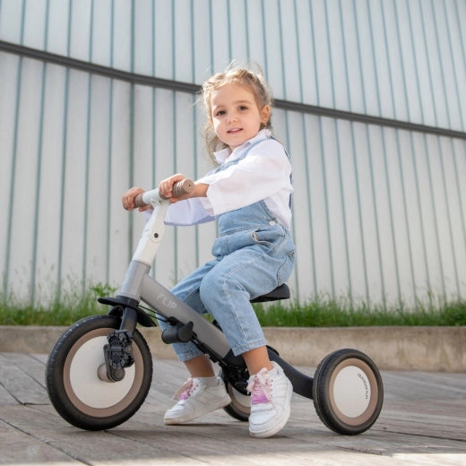 Criança sorridente a utilizar o triciclo Kikkaboo FLIP na sua configuração de bicicleta de equilíbrio/triciclo independente, em exterior com pavimento de madeira.