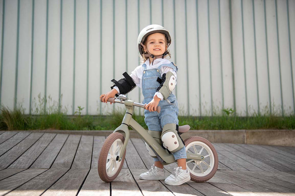 Criança sorridente pousada numa bicicleta de equilíbrio, vestindo o kit de proteções Kikkaboo (joelheiras, cotoveleiras e protetores de pulso) de cor azeitona, com capacete.
