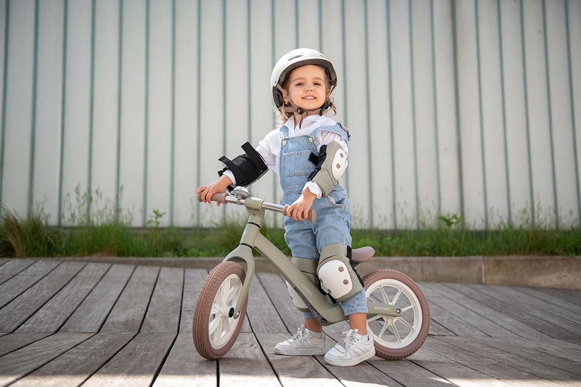 Criança sorridente pousada numa bicicleta de equilíbrio, vestindo o kit de proteções Kikkaboo (joelheiras, cotoveleiras e protetores de pulso) de cor azeitona, com capacete.