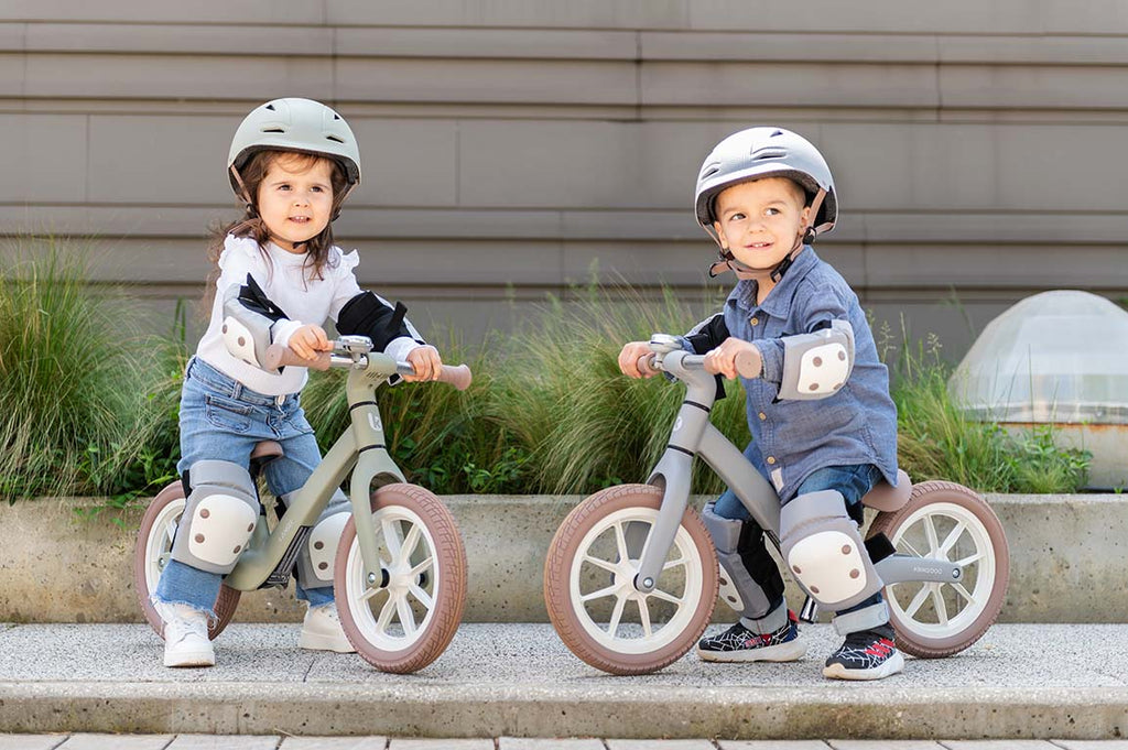 Duas crianças sorridentes, um rapaz e uma rapariga, a andar em bicicletas de equilíbrio, ambas a usar o kit de proteções Kikkaboo (joelheiras, cotoveleiras e protetores de pulso) cor de azeitona.