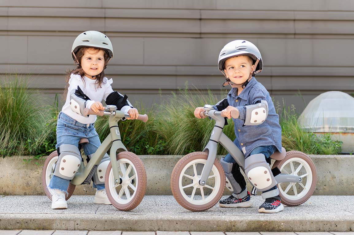 Duas crianças sorridentes, um rapaz e uma rapariga, a andar em bicicletas de equilíbrio, ambas a usar o kit de proteções Kikkaboo (joelheiras, cotoveleiras e protetores de pulso) cor de azeitona.
