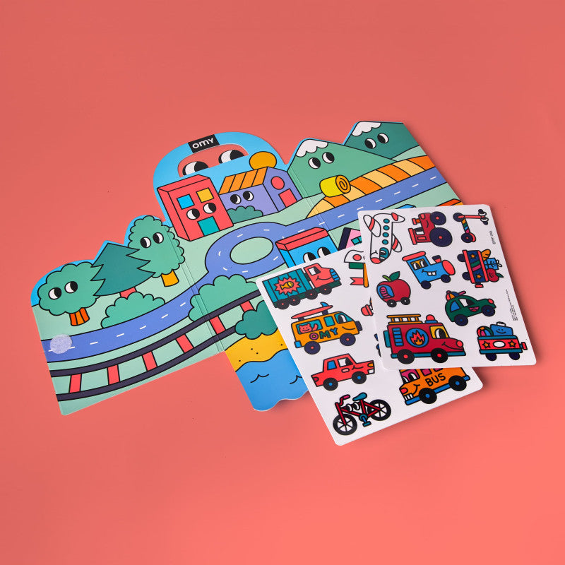 Conteúdo do Kit Montessori OMY aberto, mostrando o cenário tríptico dobrável e duas folhas de autocolantes de silicone com veículos e elementos da natureza.