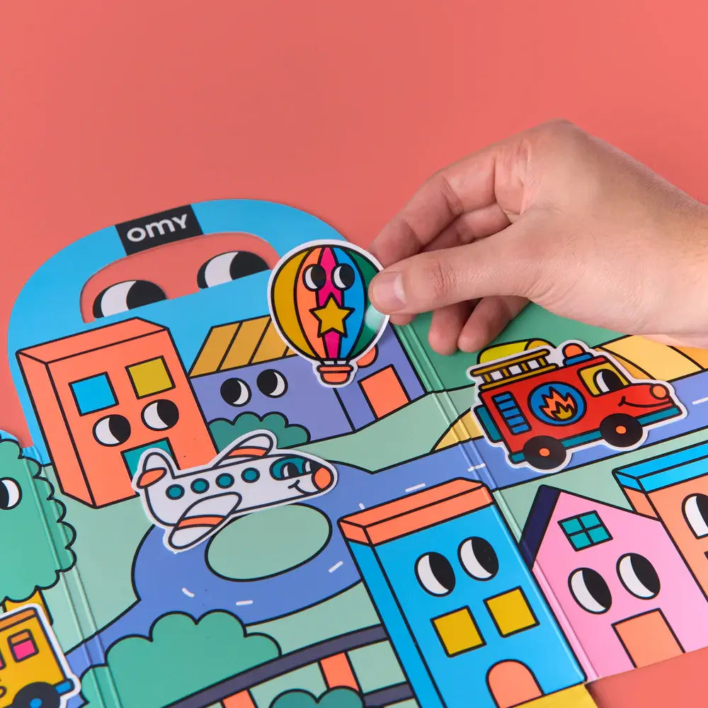 Uma mão a aplicar um autocolante de balão de ar quente colorido no cenário urbano do Kit Montessori OMY, demonstrando a facilidade de uso.