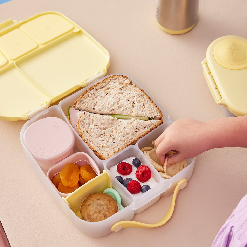 Lancheira b.box aberta mostrando uma refeição diversificada: sanduíche, iogurte com frutos vermelhos, frutos secos e batatas, com uma mão a pegar num snack.