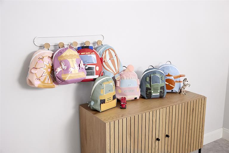 Coleção de mochilas infantis Lassig de diferentes designs (incluindo a de carrossel) penduradas num cabide e dispostas sobre um móvel de madeira clara.