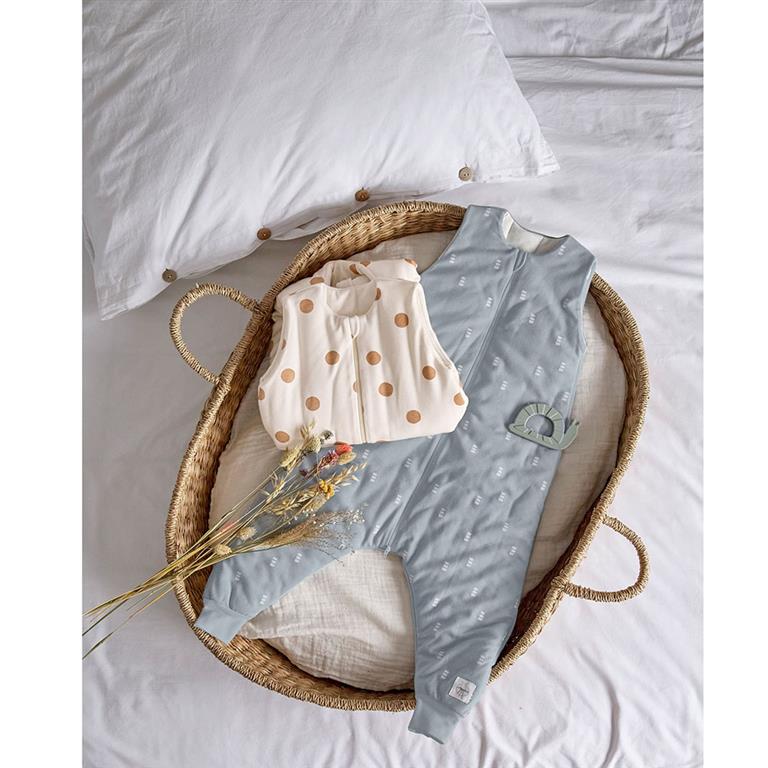 Composição de artigos Lassig num cesto de vime: saco de dormir azul com pernas e um saco de dormir bege com bolinhas, ao lado de um mordedor de caracol.