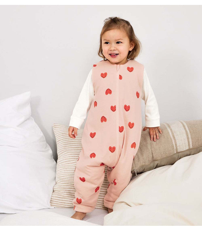 Criança pequena sorridente vestindo o saco de dormir com pernas Lassig TOG 2,5 rosa corações, em pé sobre uma cama.