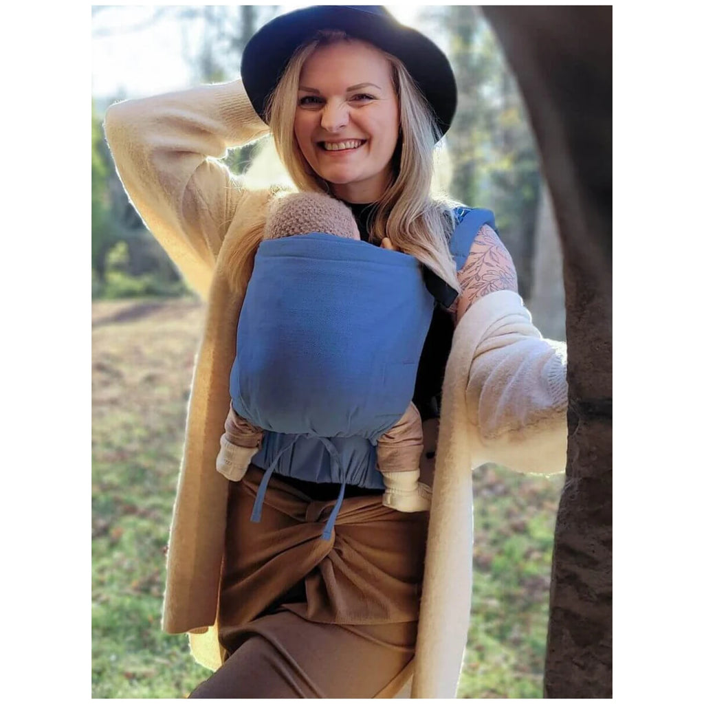 Mulher sorridente a praticar babywearing com a mochila Hoppediz Crete num ambiente exterior, destacando o conforto e proximidade.
