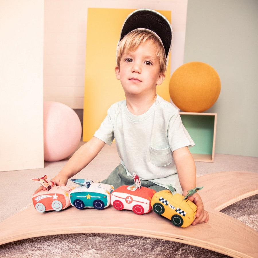 Menino brincando com 4 carrinhos de tecido Lilliputiens (taxi, polícia, ambulância, bombeiro) em tábua de equilíbrio.