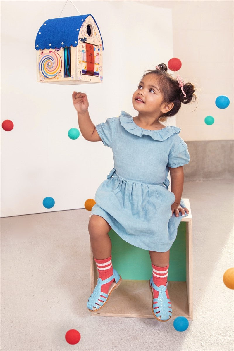 Criança pequena brinca com a Casa do Mágico Lilliputiens, um brinquedo de madeira pendurado, rodeada de bolas coloridas.