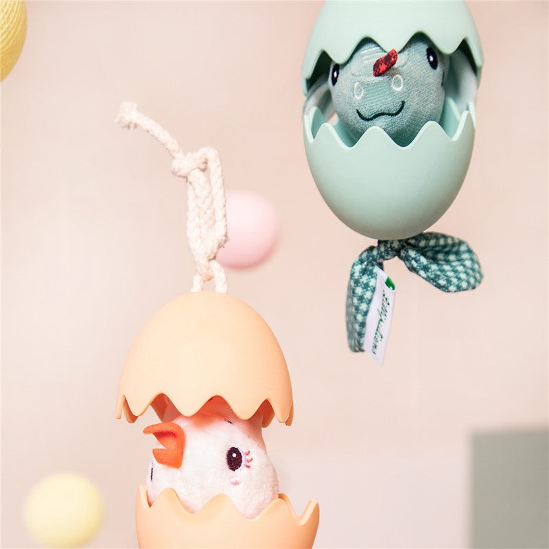 Duas figuras Lilliputiens em ovos de silicone pendurados: a galinha Paulete (pêssego) e um dinossauro (verde-água) em ambiente de berçário.