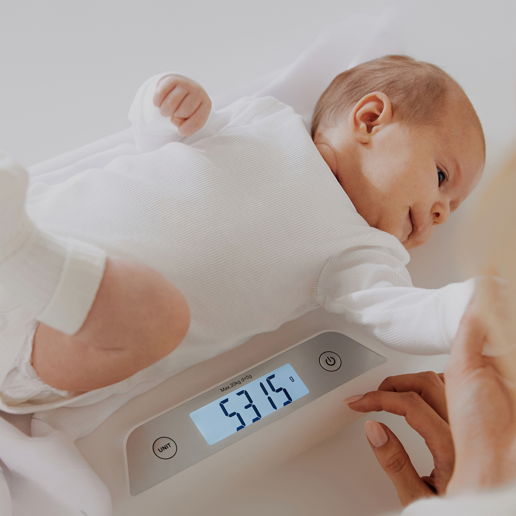 Bebé deitado confortavelmente na balança Lionelo enquanto um adulto ajusta as definições, com o peso de 5315g visível no ecrã.