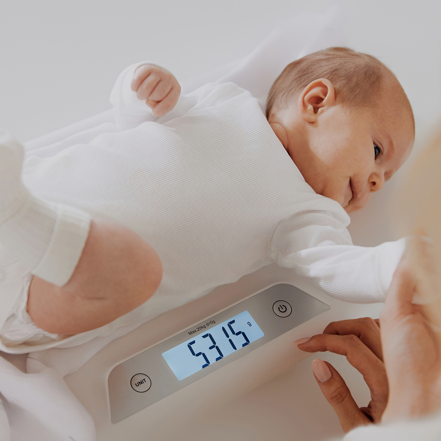 Bebé deitado confortavelmente na balança Lionelo enquanto um adulto ajusta as definições, com o peso de 5315g visível no ecrã.