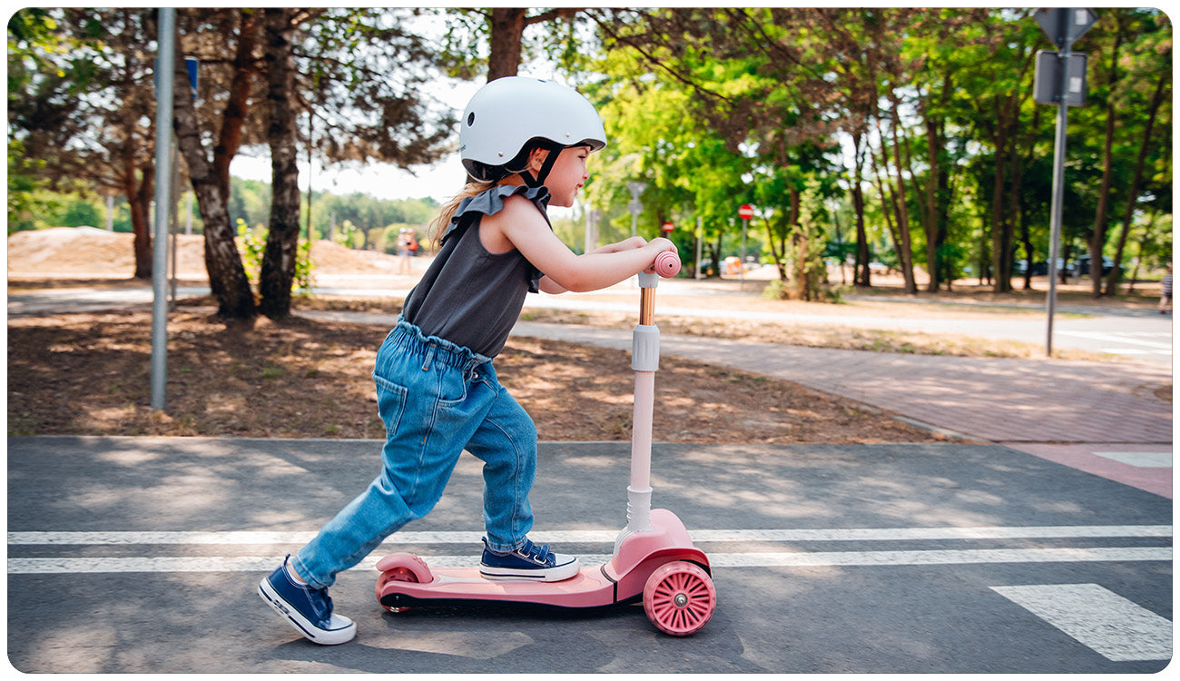 Criança a usar a trotinete de 3 rodas Lionelo Timmy cor-de-rosa num passeio, demonstrando estabilidade e diversão ao ar livre.