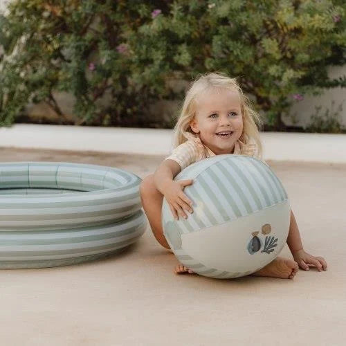 Criança loira sorridente sentada no chão ao lado de uma piscina insuflável, a segurar a bola de praia Little Dutch.