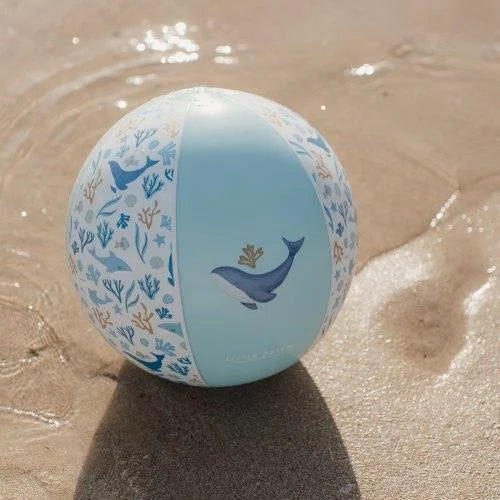 Detalhe da bola de praia Little Dutch azul com padrão de baleia e algas, pousada na areia molhada junto à água.