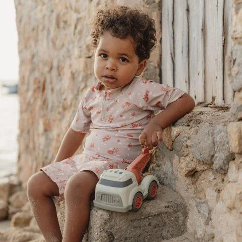 Criança pequena com fato de banho estampado a segurar o camião de reboque Little Dutch, sentada em rochas perto do mar.