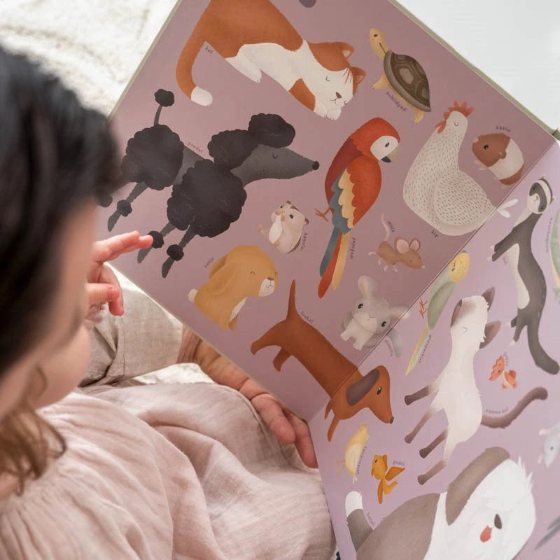 Criança a folhear o livro de cartão "My First Animal Book", mostrando uma página com ilustrações de animais domésticos como cão poodle, gato, coelho e pássaros.