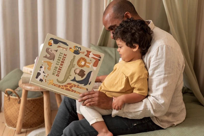 Pai a ler o livro de animais Little Dutch com o seu filho pequeno ao colo, num ambiente acolhedor com cortinas claras.