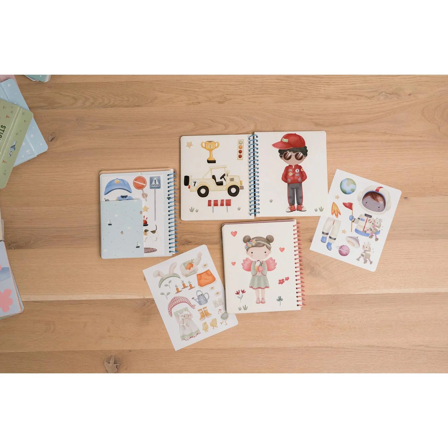 Vista aérea de vários livros de atividades Little Dutch, incluindo o Dress Me Up, abertos sobre uma superfície de madeira, mostrando diferentes temas e autocolantes.