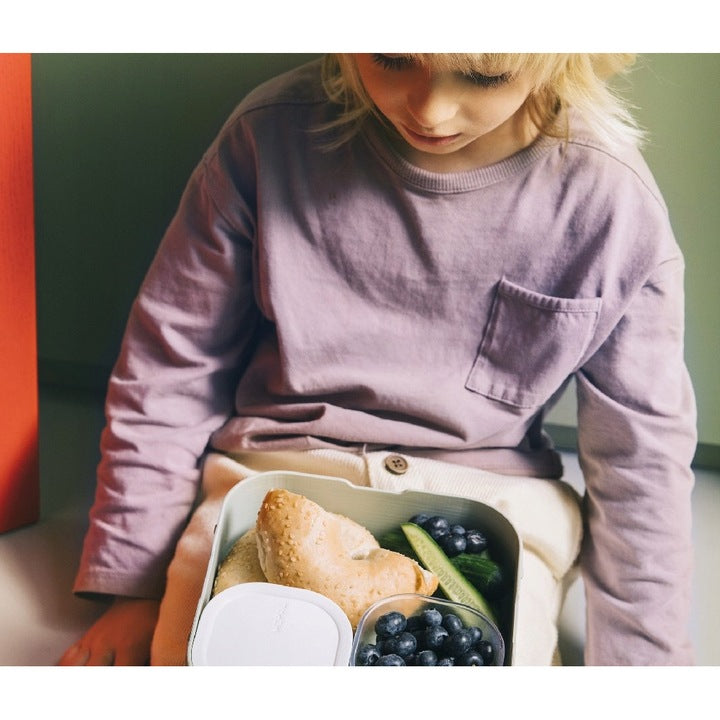 Criança loira com camisola lilás a olhar para dentro da lancheira Little Dutch cheia de pão, mirtilos e pepino, em uso prático.
