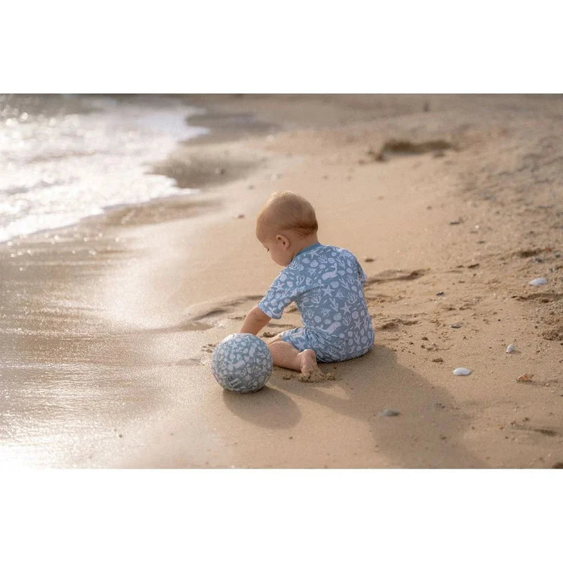 Bebé sentado na areia da praia a brincar com a Mini Bola de Praia Little Dutch com padrão marinho, perto da água.