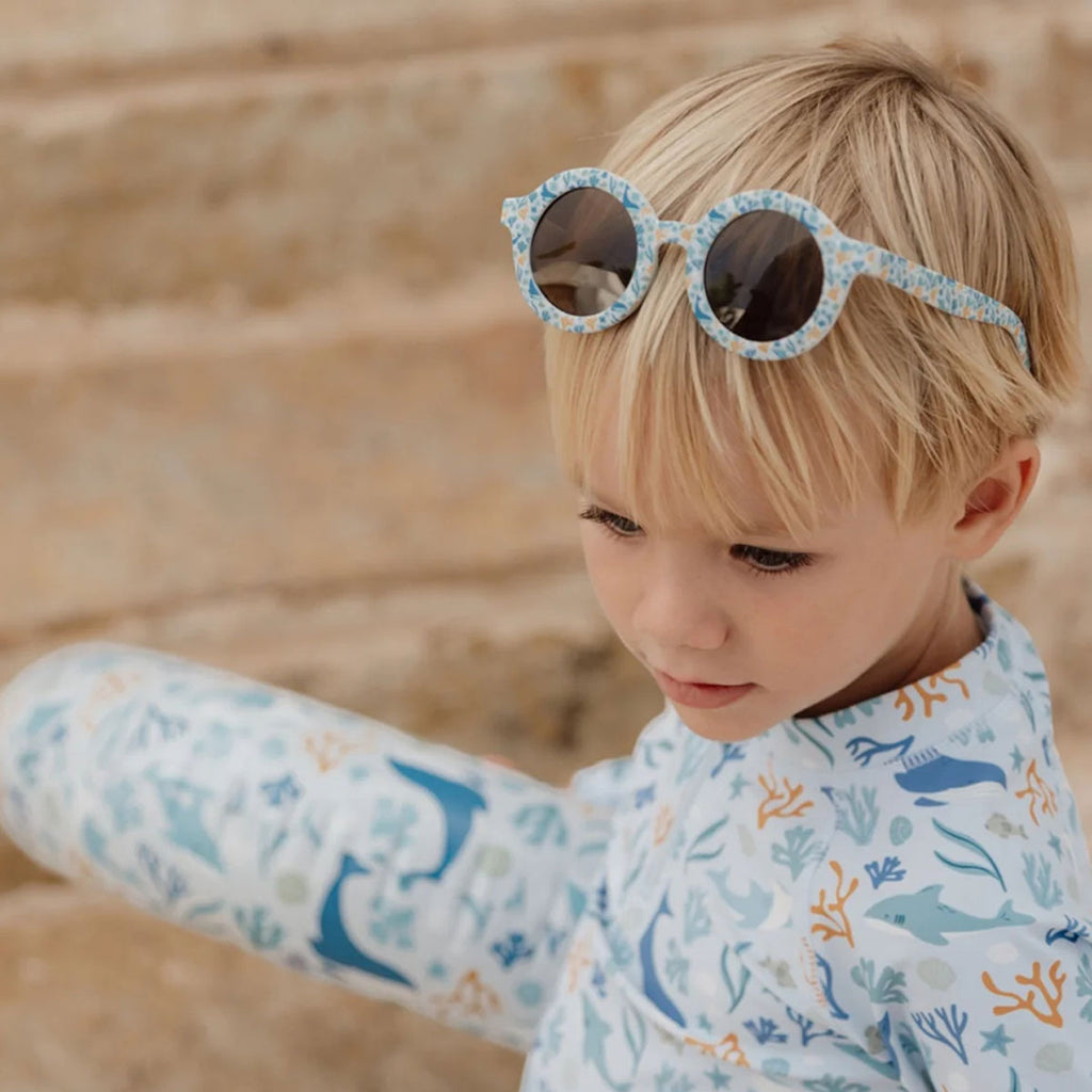 Criança loira com óculos de sol Little Dutch Ocean Dreams Blue pousados na cabeça, vestindo roupa com padrão marinho.
