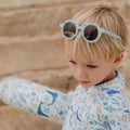 Criança loira com óculos de sol Little Dutch Ocean Dreams Blue pousados na cabeça, vestindo roupa com padrão marinho.