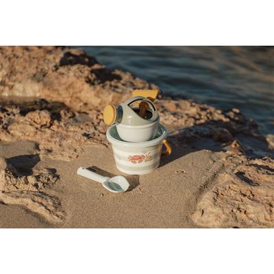 Conjunto de praia Little Dutch cinza com balde e regador pousados na areia, com a pá branca ao lado, em ambiente rochoso junto ao mar.