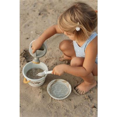 Criança a brincar na areia com o conjunto de praia Little Dutch, usando o regador para verter água num balde e a pá na mão.