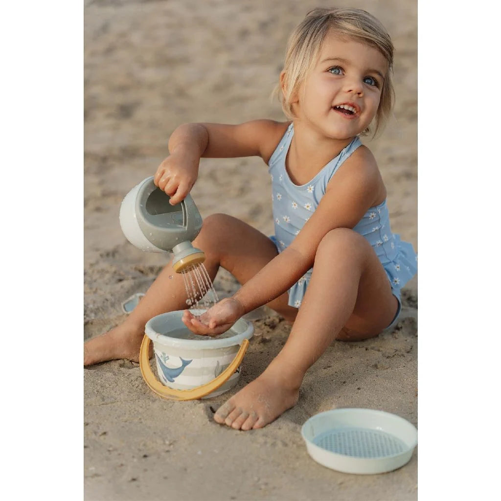 Menina sorridente na praia a usar o regador do conjunto Little Dutch para verter água na mão, com o balde e a peneira ao lado.