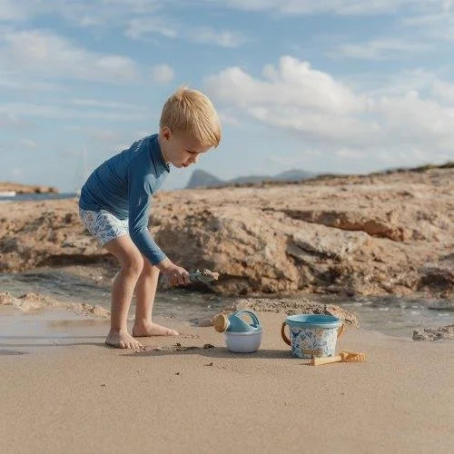 Rapaz loiro a brincar com o conjunto de praia Little Dutch (balde e regador) numa praia rochosa, com o mar ao fundo.