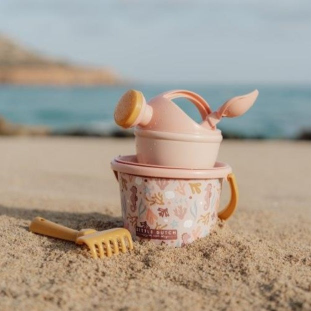Detalhe do conjunto de praia Little Dutch rosa com balde estampado de cavalos-marinhos, regador, pá e ancinho na areia junto ao mar.