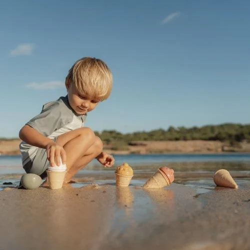 Criança loira brinca na areia com o set de gelados Little Dutch, criando formas de gelado junto à água sob o céu azul.