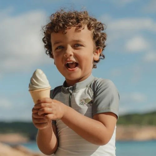 Criança com cabelo encaracolado sorri, segurando um gelado de areia feito com o set Little Dutch, em dia de sol na praia.