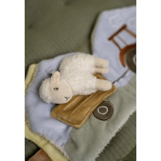 Ovelha de peluche macia deitada sobre uma página de tecido azul claro de um livro de atividades, com um trator bordado ao fundo.