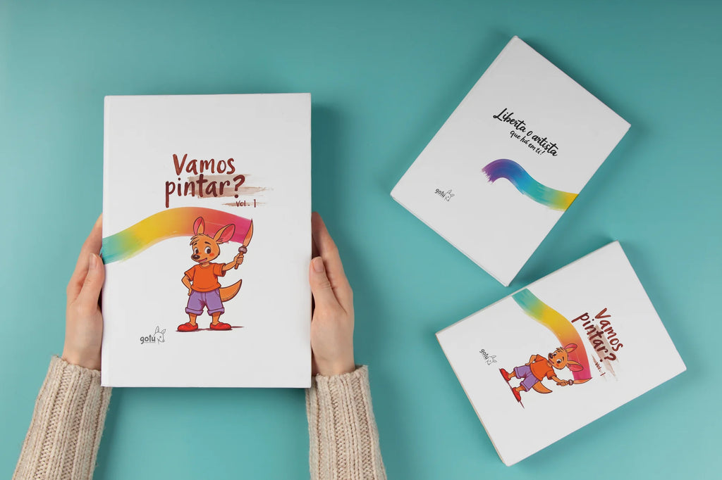 Imagem de um livro infantil para colorir intitulado "Vamos Pintar? Vol. 1", projetado para estimular a criatividade das crianças. A capa apresenta o moki segurando um pincel, com um traço colorido a acompanhar o título. No fundo, dois outros livros da mesma coleção estão sobre uma superfície azul. Um deles exibe a frase "Liberta o artista que há em ti", reforçando o tema de arte e expressão criativa. 