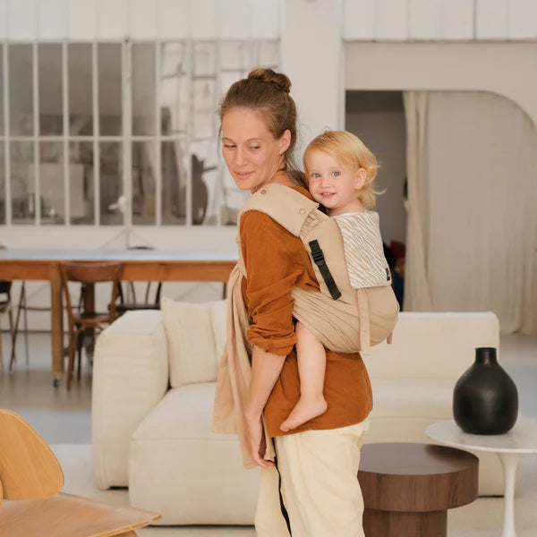 Mãe sorridente transporta uma criança pequena às costas com um porta-bebés de cor clara num ambiente doméstico moderno e luminoso.