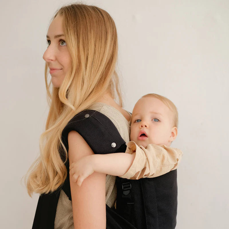 Close-up de uma mãe com cabelo loiro a olhar para o lado enquanto transporta um bebé pequeno com um porta-bebés escuro, focando-se no contacto visual e no acolchoamento da alça.