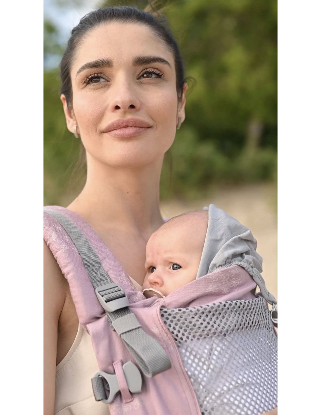 Mulher sorridente usando a mochila Love & Carry ONE - Bloom, segurando um bebé no peito, num ambiente de praia.