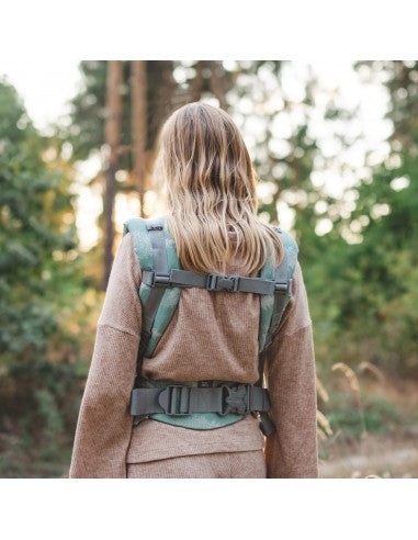 Mulher a usar a mochila Love and Carry ONE, com o bebé à frente, ao ar livre, junto com um homem ao fundo.