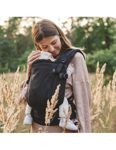 Mulher sorridente num campo de trigo, usando a Mochila Love and Carry Ajustável ONE - Twilight com o bebé adormecido no painel frontal, ambos vestidos casualmente.